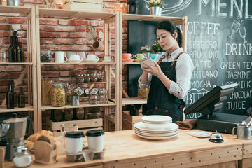 asian chinese female worker standing behind bar counter and writing notes. lady waitress work after shop close and stock taking with plates and cups in coffee shop. elegant girl staff in apron.