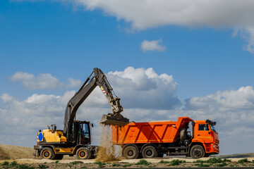 A yellow backhoe loader loads the earth into a truck during the construction of a road against a blue sky with clouds.