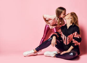 Obraz na płótnie Canvas Young beautiful red hair mother and small daughter in stylish shiny casual costumes and sunglass are posing together kissing over pink