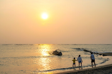Fototapeta premium View from the beach with a yellow glowing sun setting in the horizon with an Asian family playing in the water.