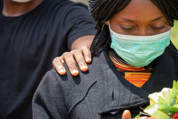a young african woman mourning, wearing black and holding flowers, someone puts a hand on her to...