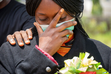 young black woman mourning, wearing black and holding flowers, someone puts a hand on her to console