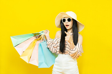 Beautiful excited happy young shopaholic asian woman wearing sungalsses and floppy hat posing isolated over yellow background holding pastel shopping bags ,summer sale concept