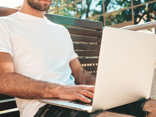 Handsome smiling stylish hipster lambersexual model. Fashion man sitting on the bench in the street and using laptop computer, writing text. Concept of co-working startup. Technology and communication