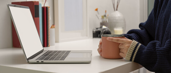 Female freelancer sitting at home office and holding coffee mug while looking on mock up laptop