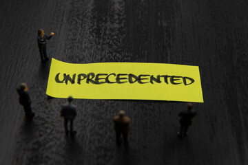 Strategy meeting with miniature figurines posed as business people standing around post-it note with Unprecedented message, minimalist abstract concept image with focus on text