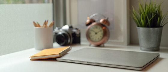 Study table with closed laptop, notebook, stationery, camera, clock and decorations on white table