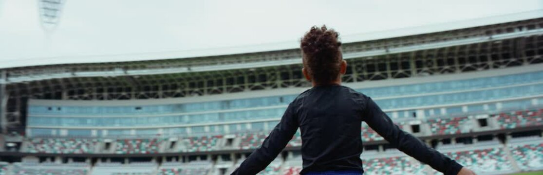 Portrait of cute little black kid boy spreading hands on a large football stadium, dreaming of becoming professional player, soccer star. Shot on RED camera with anamorphic lens