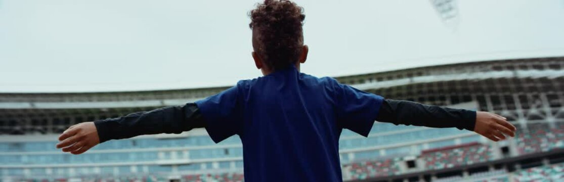 Portrait of cute little black kid boy spreading hands on a large football stadium, dreaming of becoming professional player, soccer star. Shot on RED camera with anamorphic lens
