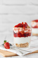 Homemade layered dessert with fresh strawberries, cream cheese or yogurt, granola and strawberry jam in glasses on white wood background. Healthy organic breakfast or snack concept. Selective focus.