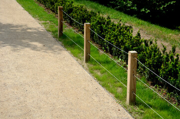 a low fence of land with three steel ropes and wooden poles separates the gravel path from the lawn and the flowerbed with boxwoods in the city park protects against entering the dogs
