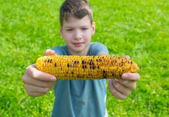close-up, a boy, against a background of green grass, shows corn cooked on the grill