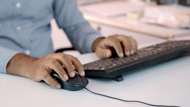 Closeup of Indian businessman typing on keyboard and scrolling mouse at desk in office