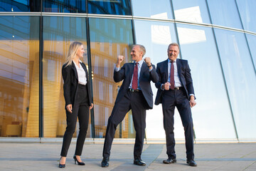 Fototapeta premium Cheerful business people celebrating victory, standing together at glass office building facade. Full length, front view. Successful team and teamwork concept