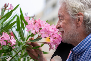 A white-haired senior man with beard lowers his medical mask to smell the pink flowers of the...