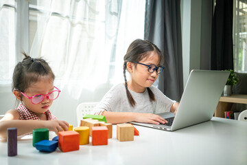 Asian girl using desktop computer for online study homeschooling during her younger sister play wooden block toy at her home. coronavirus, homeschooling or education technology concept