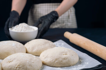 Woman in checkered apron and black gloves holds a flour bowl behind baking tray with pieces of yeast dough. Process of making bakery. Adjarian Khachapuri Recipe – Georgian cheese bread.