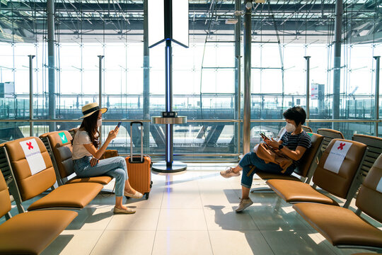 Asian female tourist wearing mask using mobile phone searching airline flight status and sit social distancing chair in airport during coronavirus or covid-19 virus outbreak a new normal concept