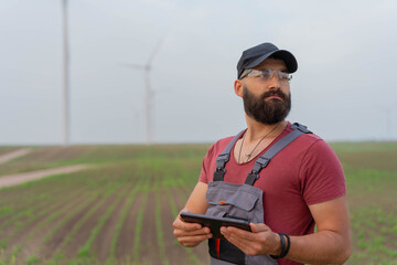 Young worker with strong beard in wind park. Holding tablet. Wind Mill  in background.