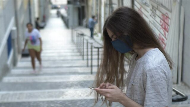 Long Haired Face Masked Guy Using Smartphone Outside. Young man with long hair wearing a face mask protection while using a smartphone in a big stairway in Lisbon, Portugal