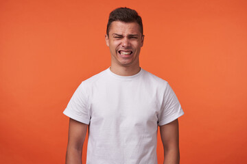 Portrait of young brunette male with short haircut grimacing his face and showing teeth while looking at camera, standing over orange background with hands down