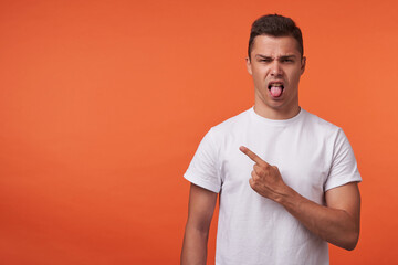 Displeased young short haired brunette man frowning his face and sticking out tongue while pointing aside with forefinger, isolated over orange background