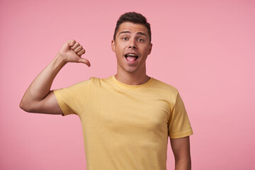Indoor photo of young pretty brown haired guy with short haircut thumbing on himself while looking excitely at camera with opened mouth, posing over pink background