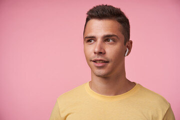 Portrait of positive young handsome short haired brunette male with earpiece looking wonderingly aside while standing over pink background with hands down