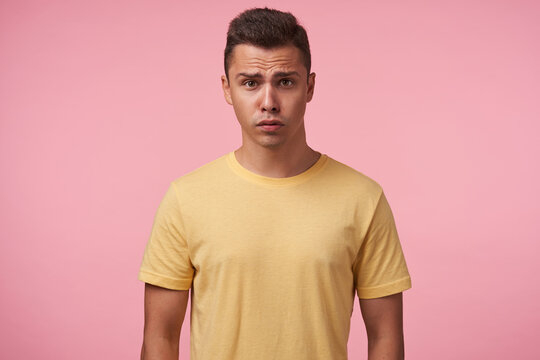 Bewildered young handsome short haired brunette man grimacing his face while looking confusedly at camera, keeping hands down while posing over pink background