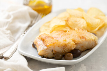 fried cod fish with olives and potato chips on white dish on ceramic background