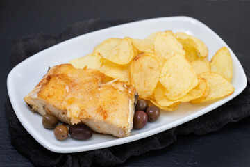 fried cod fish with olives and potato chips on white dish on ceramic background