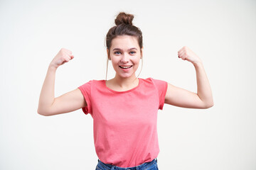 Fototapeta na wymiar Cheerful young attractive brown haired woman with natural makeup raising her hands while showing biceps and smiling gladly at camera, isolated over white background