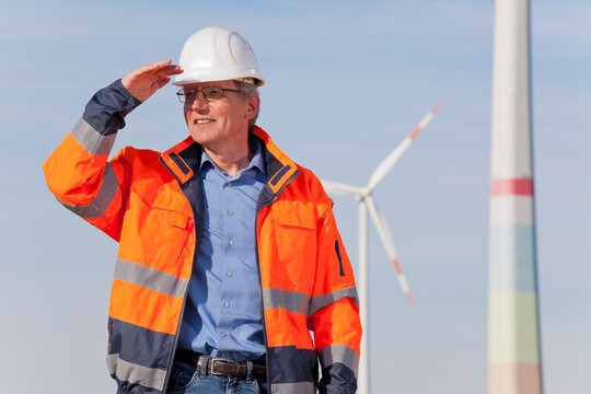 Engineer with hard hat and protective clothing in front of a windfarm looking for a bright future for green energy