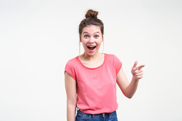 Fototapeta na wymiar Excited young pretty dark haired female with natural makeup looking amazedly at camera with wide mouth opened while showing aside with forefinger, isolated over white background