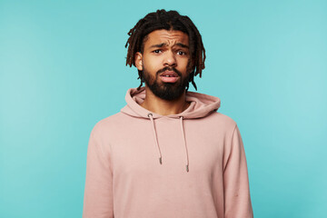 Bewildered young brown-eyed brunette bearded male frowning his eyebrows while looking confusedly ar camera, isolated over blue background in sporty wear