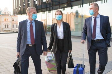 Business tourists in face masks walking outdoors with luggage, talking to each other. Front view....