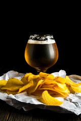 A glass of beer and potato chips on a wooden table and a dark background