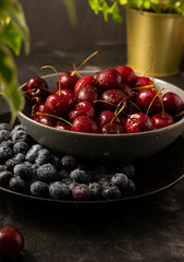 Selective focus, ripe cherries and blue blueberries with water drops in a dark kitchen dish, on a dark background, vertical position, rustic style