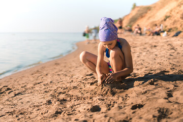 A little girl child in a swimsuit and a bandana protecting her from the sun sits on the sand on the beach and plays with the sand, builds castles, a place for text, the concept of a summer vacation
