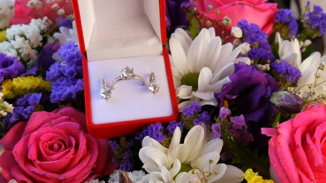 Gift present red box with a set of engagement ring and earrings on the background of a multi-colored beautiful bouquet of flowers. Marriage proposal. White gold or silver with diamond stone