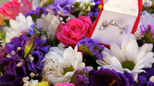 Gift present red box with a set of engagement ring and earrings on the background of a multi-colored beautiful bouquet of flowers. Marriage proposal. White gold or silver with diamond stone