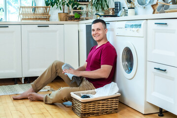 young man is sitting at home in the kitchen near the washing machine and smiling. The concept of washing is easy.