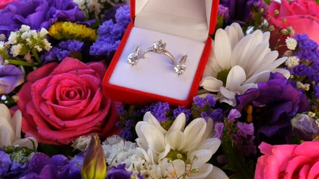 Gift present red box with a set of engagement ring and earrings on the background of a multi-colored beautiful bouquet of flowers. Marriage proposal. White gold or silver with diamond stone