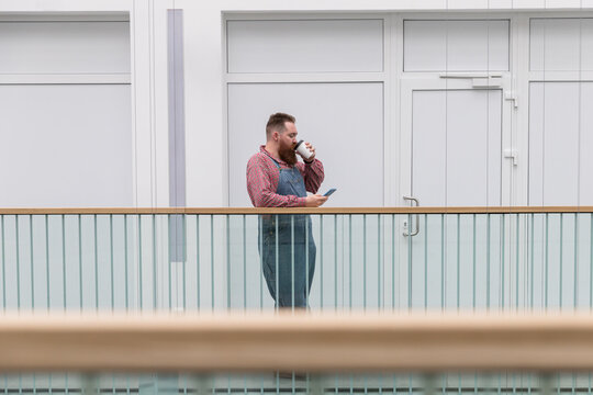 Brutal bearded self employed worker in blue overalls, checked shirt, drinking coffee from a paper cup, using smartphone. Time to take break. Hipster barber man resting. A break during work