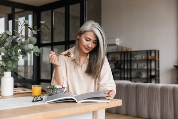 Woman sitting at kitchen reading book and eating salad.