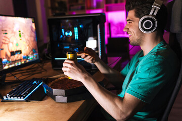 Image of joyful young man eating pizza while playing video game