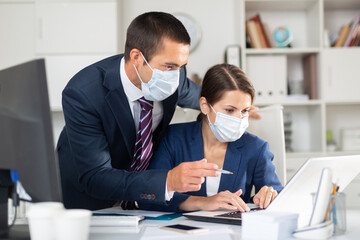 Portrait of two office employees in medical masks concentrating on work with papers and laptop. Necessary precautions during coronavirus pandemic