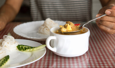 Hand with silver sppon scooping Thai curry in white soup bowl in restaurant