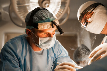 2 surgeons with headlamp do plastic operation in medical clinic. Chest augmentation plastic operation and correction in medical clinic. Group of professional medics in medical masks do plastic surgery