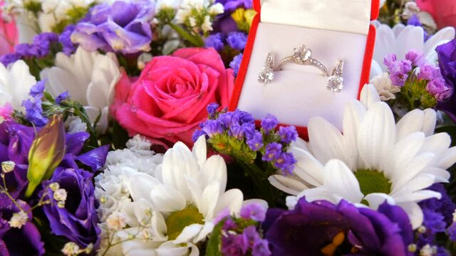 Gift present red box with a set of engagement ring and earrings on the background of a multi-colored beautiful bouquet of flowers. Marriage proposal. White gold or silver with diamond stone